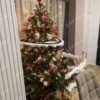 Umělý vánoční stromek 3D Smrk Skandinávský 180cm je vkusně vyzdoben bílými a červenými dekoracemi a noční vlakovou výzdobou s kolejnicemi.