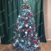 Umělý vánoční stromeček s tmavozelenými větvičkami, ozdobený červenými a zelenými ozdobami a bílým osvětlením, v obýváku