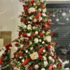 Umělý vánoční stromeček se zelenými větvičkami, hustě ozdobený červeno-bílými ozdobami a teplým bílým osvětlením, v rohu obývacího pokoje