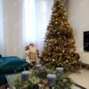Široký a hustý umělý vánoční stromeček s bledě zelenými hustými větvičkami, hustě ozdobený měděno-zlatými ozdobami a teplým bílým osvětlením, v rohu obývacího pokoje