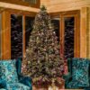 Vysoký a široký umělý vánoční stromeček s bledě zelenými hustými větvičkami, hustě ozdobený fialovými ozdobami a teplým bílým osvětlením, v rohu obývacího pokoje