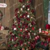 Široký a hustý umělý vánoční stromeček s bledě zelenými hustými větvičkami, hustě ozdobený červenými ozdobami, v rohu obývacího pokoje