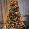 Umělý vánoční stromeček se zasněženými konci větviček a šiškami, ozdobený červenými lesními plody, hustě ozdobený červeno-zlatými ozdobami a teplým bílým osvětlením, v rohu obývacího pokoje