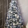 Široký a vysoký umělý vánoční stromek se zasněženými konci větviček, hustě ozdobený stříbrno - bílými ozdobami, v rohu obývacího pokoje
