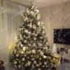 Vysoký a široký umělý vánoční stromeček se zelenými větvičkami, ozdobený bílo-stříbrnými ozdobami, s teplým bílým osvětlením, v rohu obývacího pokoje