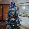 Umělý vánoční stromeček s hustými zelenými větvičkami, ozdobený bílo-modrými ozdobami a bílým osvětlením, v obýváku