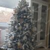 Bílý umělý vánoční stromeček, s hustě zasněženými větvičkami, ozdobený bílými a zlatými ozdobami, u okna v obýváku