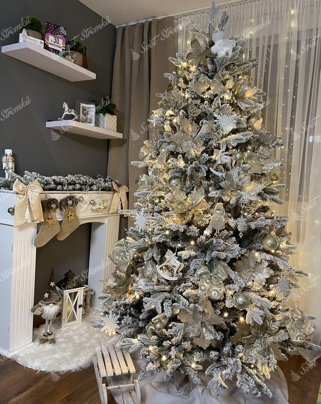 Umělý vánoční stromeček se zasněženými větvičkami, ozdobený v glamour stylu bílo-zlatými ozdobami, s teplým bílým osvětlením, v rohu obývacího pokoje u krbu