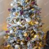 Bílý hustý umělý vánoční stromek se zasněženými větvičkami, elegantně ozdobený růžovo-bílými ozdobami, s teplým bílým osvětlením, v obýváku