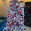 Zasněžený umělý vánoční stromeček s hustými větvičkami, hustě ozdobený bílo-červenými ozdobami, s bílým studeným osvětlením, v obýváku
