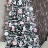Bílý umělý vánoční stromeček s hustě zasněženými větvičkami, ozdobený bílo-růžovými ozdobami, v rohu obývacího pokoje