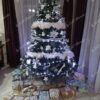 Vysoký a mohutný umělý vánoční stromeček se zelenými větvičkami, ozdobený bílými ozdobami s bílým osvětlením, v rohu obývacího pokoje