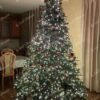 Vysoký a široký umělý vánoční stromeček se zelenými větvičkami, elegantně a hustě ozdobený červeno-zlatými ozdobami s bílým osvětlením, ve velkém obývacím pokoji