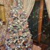 Vysoký bílý umělý vánoční stromeček s hustě zasněženými větvičkami, elegantně a hustě ozdobený bílo-růžovými ozdobami, v rohu obývacího pokoje u okna