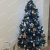 Umělý tmavě zelený vánoční stromek s postříbřenými konci větviček, ozdobený zlatými a přírodními ozdobami, s bílým kobercem pod stromeček, v rohu obývacího pokoje