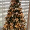 Umělý vánoční stromeček s bledě zelenými větvičkami, ozdobený bílými ozdobami, hvězdičkami a mašlemi, s teplým bílým osvětlením, u okna v obýváku