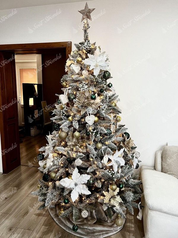 Umělý vánoční stromek se zasněženými větvičkami, ozdobený zlato-bílými ozdobami a vánočními růžemi, s teplým bílým osvětlením, v obýváku