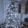 Zasněžený vysoký umělý vánoční stromeček, ozdobený růžovo-bílými ozdobami, se studeným bílým osvětlením, v obýváku