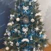 Umělý vánoční stromek s ledově zelenými větvičkami, ozdobený bílo-stříbrnými ozdobami, s kobercem pod stromeček v obýváku