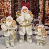 Vánoční dekorace Santa Claus Bílo Zlatý, má na sobě bílo-zlatý kabát s bílou kožešinou a v ruce plyšovou hračku.